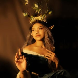 May include: A woman wearing a dark green velvet off-the-shoulder dress with a white trim. She has long, gray hair and is adorned with a floral and butterfly crown. The image has a warm, golden lighting.