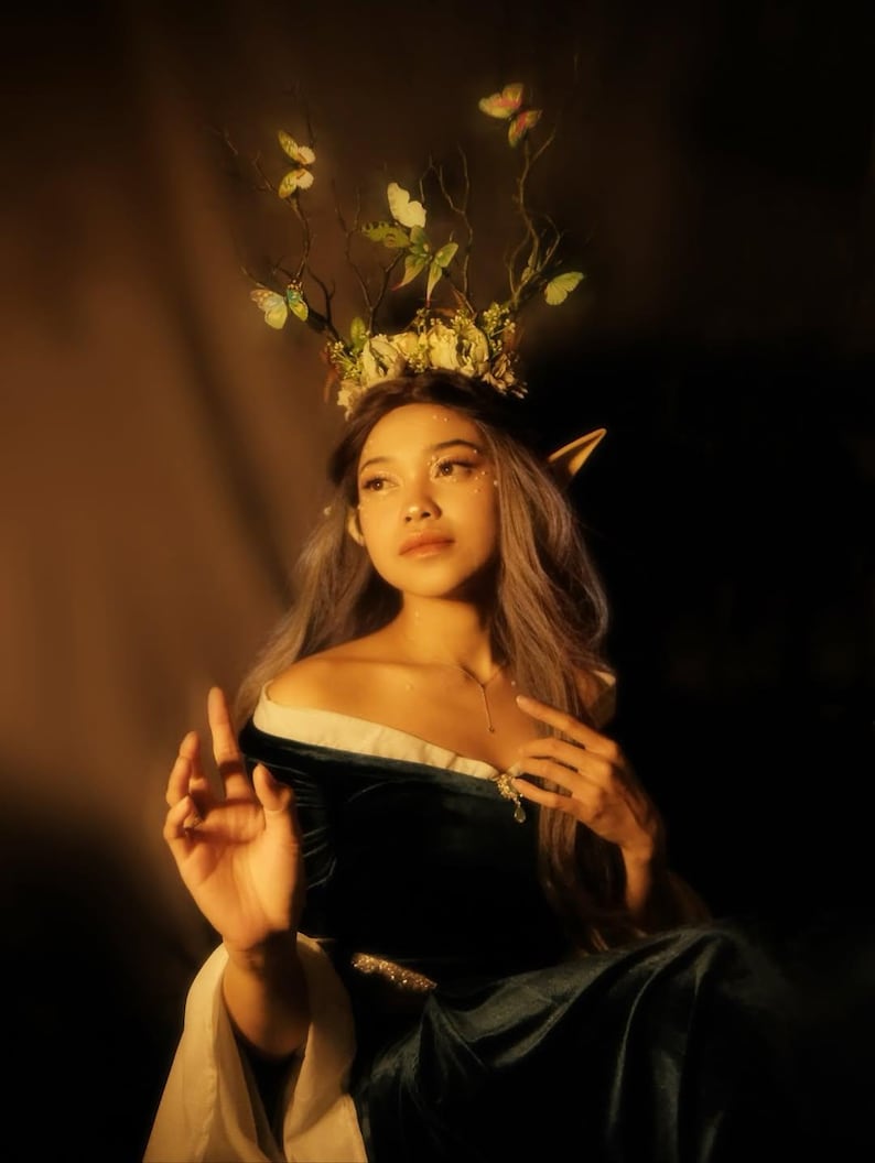 May include: A woman wearing a dark green velvet off-the-shoulder dress with a white trim. She has long, gray hair and is adorned with a floral and butterfly crown. The image has a warm, golden lighting.
