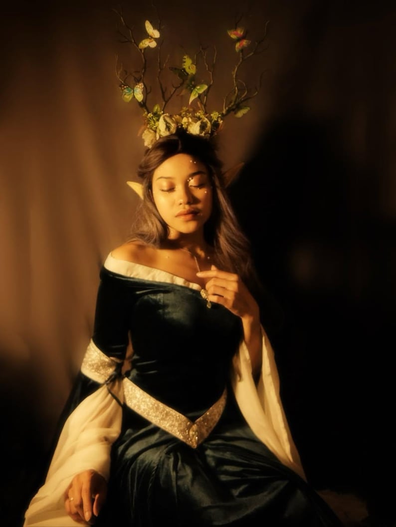 May include: A woman wearing a dark teal velvet off-the-shoulder dress with white sleeves and a gold embellished belt. She wears a headpiece of branches, flowers, and butterflies, and has pointed ears. The overall theme is fantasy.
