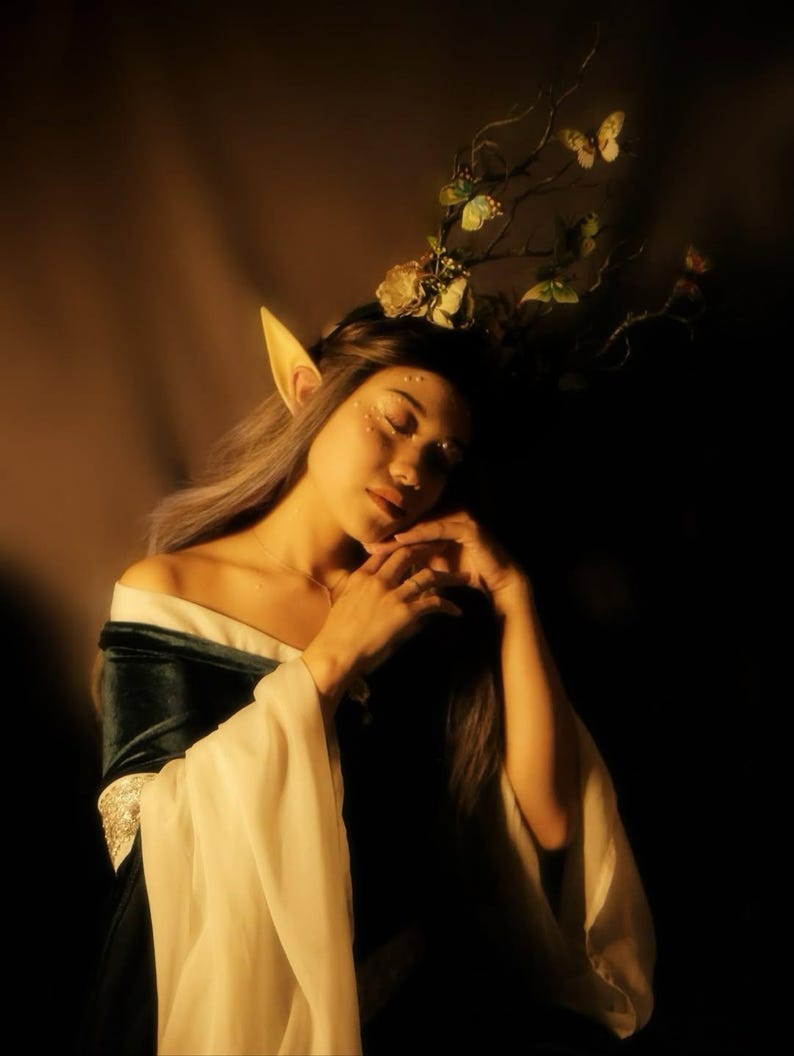 May include: A person wearing a dark green and white off-the-shoulder dress with long, flowing sleeves. The person has long, silver hair, pointed ears, and a floral and butterfly headpiece. The person is wearing a necklace and has glitter on their face.