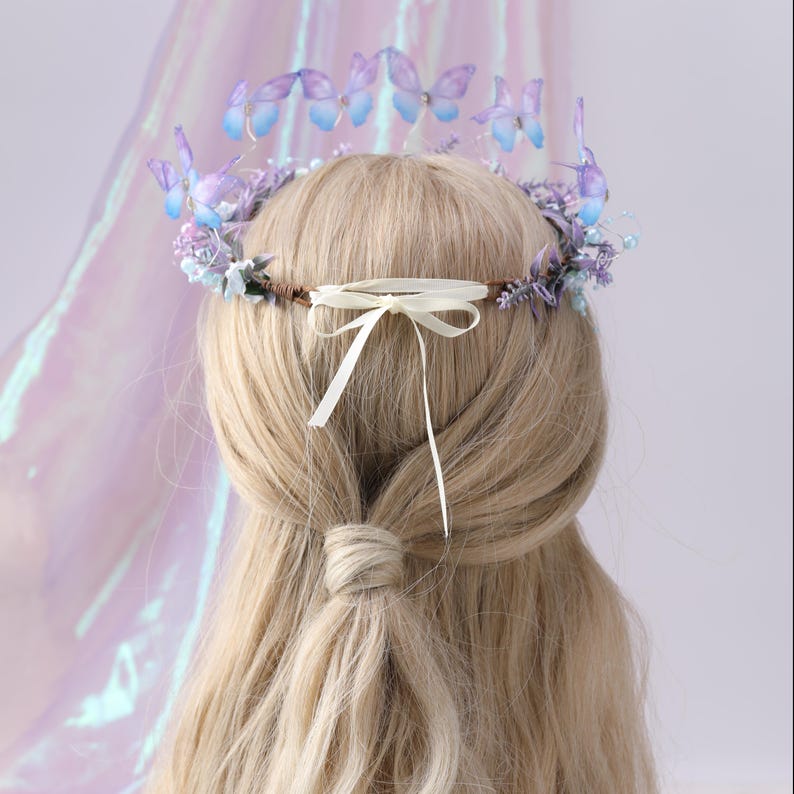 May include: A delicate butterfly crown with purple and blue butterflies, small flowers, and pearls. The crown is attached to a brown band and tied with a cream-colored ribbon. The hair is long and blonde.