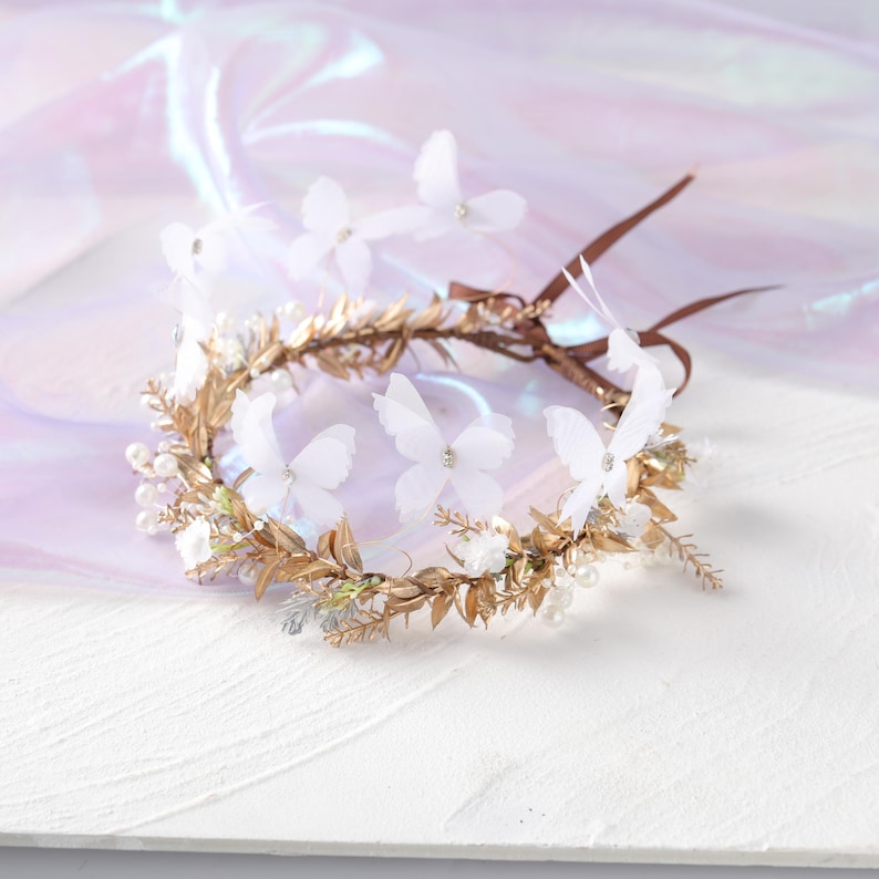May include: A gold-toned floral crown with white butterfly accents and small pearls. The crown features delicate gold leaves and a brown ribbon for securing. The background is a soft, iridescent fabric.