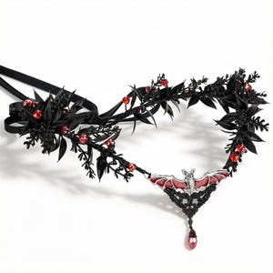 May include: A headpiece featuring a central silver-toned bat pendant with red enamel wings, accented by black faux foliage and small red beads. The structure is connected to black satin ribbons for tying, creating a decorative accessory with a dark aesthetic.