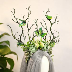 May include: A whimsical headpiece featuring faux branches, green butterflies, and floral accents. The crown is adorned with various shades of green and white flowers, creating a natural and enchanting aesthetic. The headpiece is designed to be worn on the head.