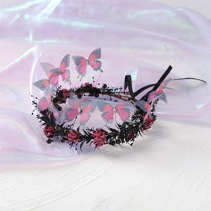 May include: A floral crown featuring red roses, black leaves, and delicate pink butterflies. The butterflies have a gradient effect, transitioning from pink to translucent. Black ribbon ties complete the design, perfect for a special occasion.