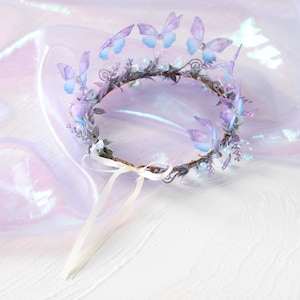 May include: A delicate, handcrafted crown featuring a circular base adorned with purple and blue butterflies. The crown is embellished with small white flowers, sprigs of greenery, and faux pearls. A white ribbon is attached for securing the crown.