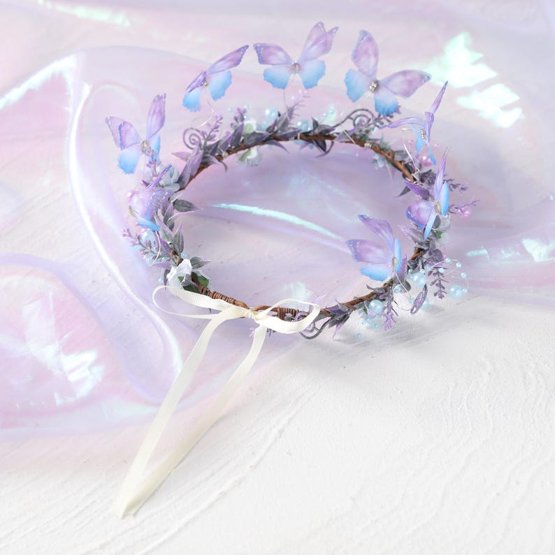 May include: A delicate, handcrafted crown featuring a circular base adorned with purple and blue butterflies. The crown is embellished with small white flowers, sprigs of greenery, and faux pearls. A white ribbon is attached for securing the crown.