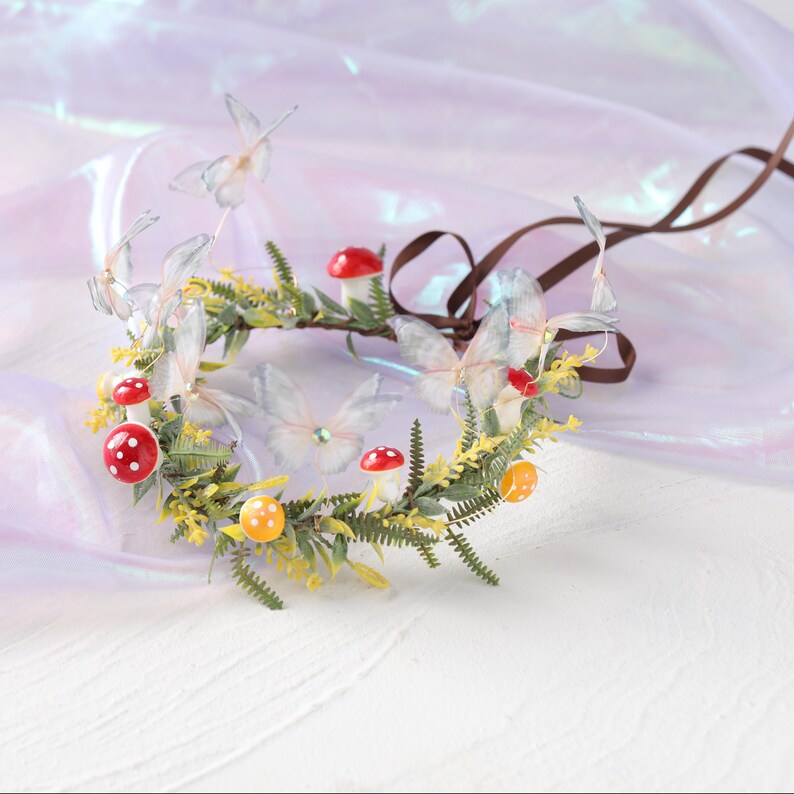 May include: A whimsical floral crown featuring delicate, translucent butterflies, red and yellow spotted mushrooms, and green foliage. The crown is adorned with a brown ribbon for tying. Perfect for fairy-themed events or costume parties.