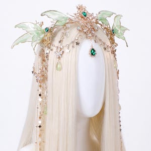 May include: A gold-toned headpiece featuring translucent light green butterfly wings, clear crystals, and emerald-coloured teardrop gems. The accessory includes delicate hanging chains with small butterfly charms and beads that drape down the sides.