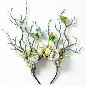 May include: A whimsical headband featuring faux branches, white and green flowers, and colorful butterfly accents. The dark green branches extend upwards, with the butterflies perched on top. The headband is black.