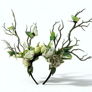 May include: A whimsical headpiece featuring a black headband adorned with faux branches, green butterflies, and cream-colored flowers. The branches extend upwards, creating a unique, nature-inspired design. The butterflies add a touch of color and fantasy.