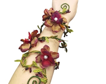 May include: A flexible arm cuff featuring a winding green vine structure adorned with deep red and brown fabric flowers. Each flower is centered with a small white pearl bead. The design includes small green leaves and dark red berry accents.