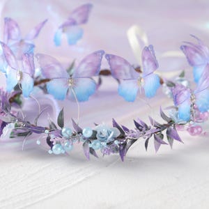 May include: A delicate floral crown featuring lavender and blue butterfly accents. The crown is adorned with small blue flowers, pearls, and purple leaves. The butterflies have a gradient of purple and blue, with a small jewel in the center.