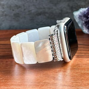 May include: A silver Apple Watch with a white, rectangular, mother-of-pearl watch band. The band is made of several rectangular pieces. The watch face is black. The watch is on a wooden surface.