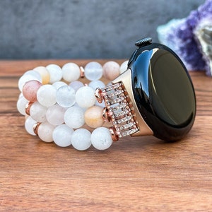 May include: A rose gold smartwatch with a black face and a white beaded band. The band is made of multiple strands of white, pink, and beige beads.