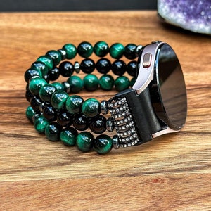Puede incluir: Una pulsera de reloj de cuentas negras y verdes con una esfera de reloj plateada. La pulsera est&aacute; hecha de m&uacute;ltiples hileras de cuentas y tiene un cierre de metal negro.