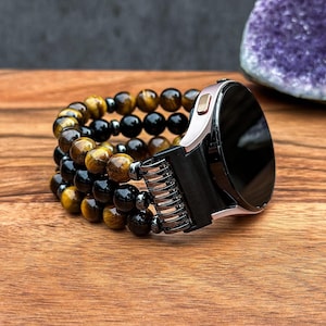 May include: A smartwatch with a black screen and silver casing is attached to a beaded bracelet. The bracelet features alternating rows of tiger's eye, black onyx, and hematite beads. The watch and bracelet rest on a wooden surface.