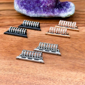 May include: Five different Apple Watch band connectors with rows of clear rhinestones. The connectors are in black, silver, and rose gold.