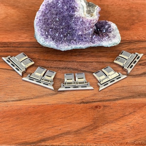 May include: Silver and gold watch band adapters are arranged on a wooden surface, with a large purple amethyst geode in the background. The adapters have a rectangular design with a silver base and gold accents.