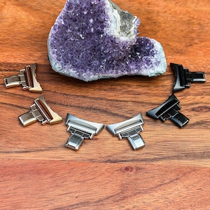 May include: A selection of watch clasps in silver, gold, and black finishes are displayed on a wooden surface. A large, raw amethyst geode is in the background. The clasps are designed for watch straps and are made of metal.