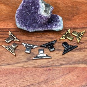 May include: Various watch strap adapters in gold, silver, and black, displayed on a wooden surface. A large, purple geode crystal is in the background. The adapters are designed to connect watch straps to the watch face.