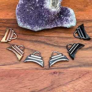 May include: Assortment of metal purse hardware in gold, rose gold, silver, and black finishes. The hardware features a curved base with a rectangular bar and a D-ring. A large purple amethyst geode is in the background.