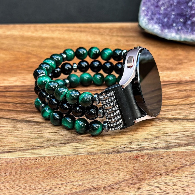 Puede incluir: Una pulsera de reloj de cuentas negras y verdes con una esfera de reloj plateada. La pulsera est&aacute; hecha de m&uacute;ltiples hileras de cuentas y tiene un cierre de metal negro.