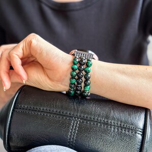 Puede incluir: Una pulsera de reloj negra y verde con cuentas y un cierre plateado. La pulsera est&aacute; hecha de m&uacute;ltiples hileras de cuentas, con algunas cuentas negras y algunas cuentas verdes.