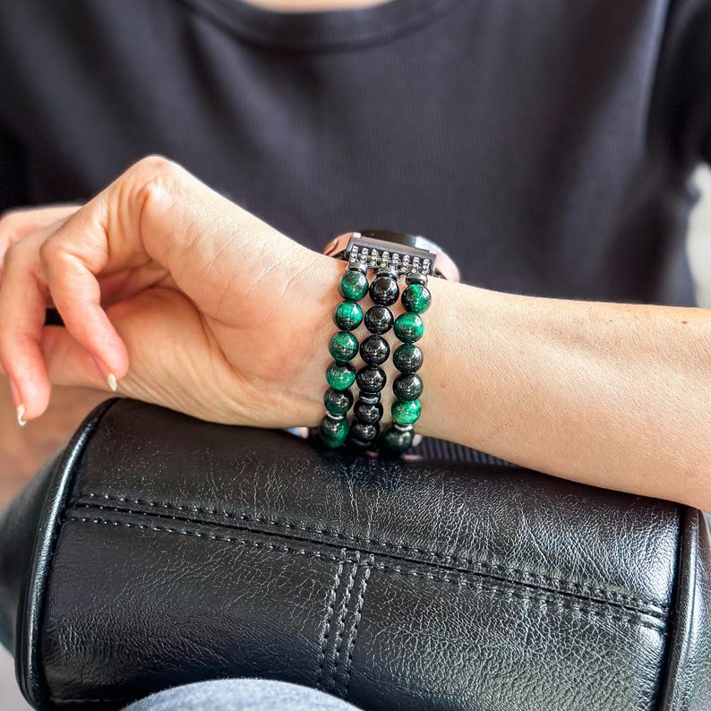 Puede incluir: Una pulsera de reloj negra y verde con cuentas y un cierre plateado. La pulsera est&aacute; hecha de m&uacute;ltiples hileras de cuentas, con algunas cuentas negras y algunas cuentas verdes.