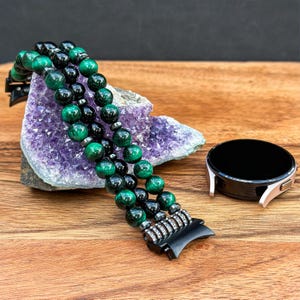 Puede incluir: Una pulsera de reloj de cuentas negras y verdes con un cierre de metal. La pulsera est&aacute; hecha de m&uacute;ltiples hilos de cuentas y descansa sobre una geoda de amatista p&uacute;rpura.