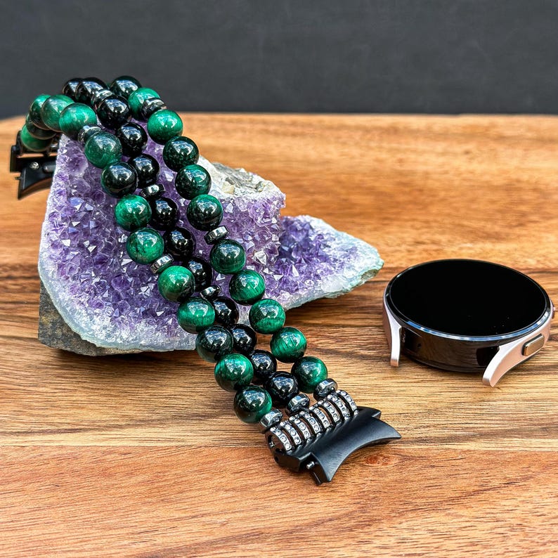 Puede incluir: Una pulsera de reloj de cuentas negras y verdes con un cierre de metal. La pulsera est&aacute; hecha de m&uacute;ltiples hilos de cuentas y descansa sobre una geoda de amatista p&uacute;rpura.