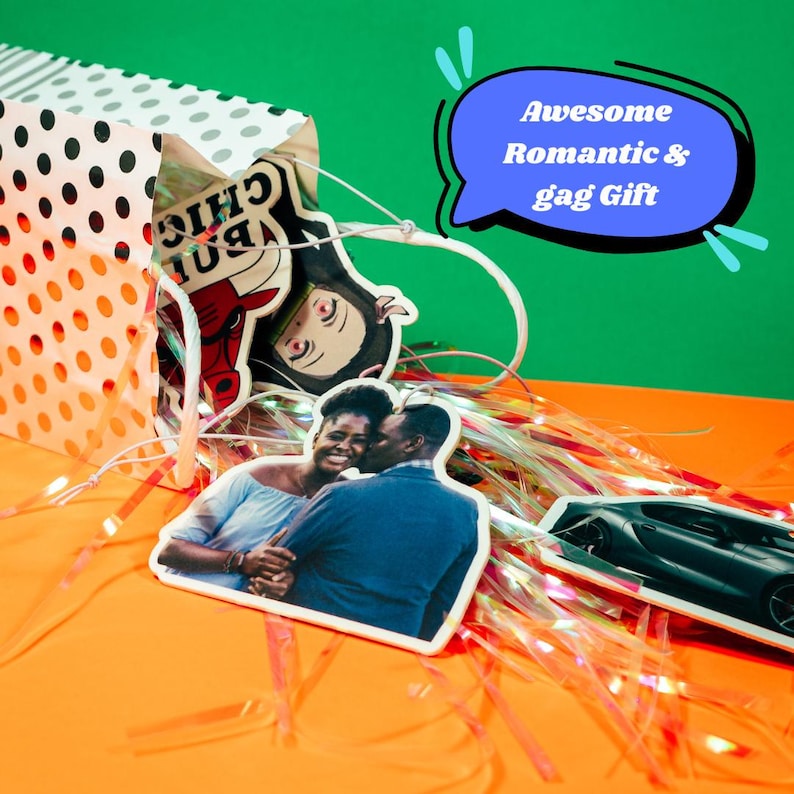 May include: A white paper bag with black polka dots filled with various car air fresheners. The air fresheners include a cartoon image of a bull, a cartoon image of a person, a photo of a couple, and a photo of a car. The bag is sitting on a bright orange surface with pink and white iridescent streamers.