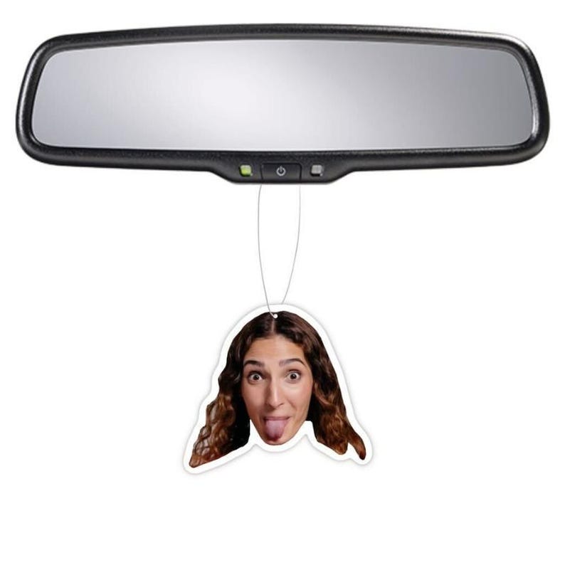 May include: A car air freshener featuring a woman's face. The woman has brown hair and is sticking her tongue out.