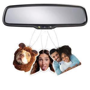 May include: Three car air fresheners hanging from a rear view mirror. One features a dog wearing a brown bear costume, another features a woman with her tongue sticking out, and the last one features a couple hugging.