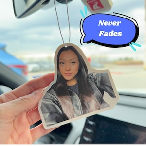 May include: A white car air freshener featuring a photo of a person with long dark hair wearing a black leather jacket. The air freshener has a blue speech bubble with the text "Never Fades" on it.