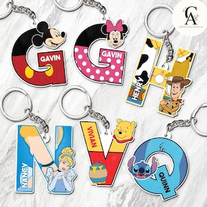 May include: A collection of colorful character-themed keychains. The keychains feature the letters G, H, N, V, and Q, each with a different character design, including Mickey Mouse, Minnie Mouse, Woody, Cinderella, Winnie the Pooh, and Stitch. Each keychain has a name.