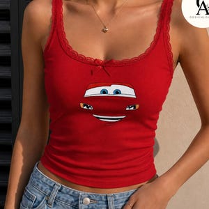 May include: A bright red ribbed tank top featuring a graphic of a car face with blue eyes and a wide smile. The neckline and straps are trimmed with delicate red lace, and a small red ribbon bow is centered at the top of the chest area.