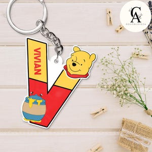 May include: A keychain featuring the letter "V" in red and yellow, with the name "VIVIAN" printed vertically. It includes a cartoon image of Winnie the Pooh and a honey pot. The keychain is attached to a silver chain and ring.