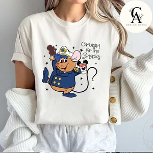 May include: White t-shirt with a cartoon mouse wearing a captain's hat and holding a Mickey Mouse ice cream cone and a cup of coffee. The text "Cruisin' for the Snacks" is printed on the shirt.