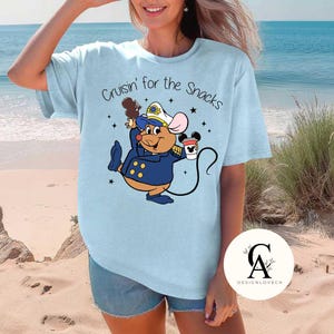 May include: A light blue t-shirt with a cartoon mouse wearing a captain's hat and holding a cup of coffee. The text "Cruisin' for the Snacks" is printed on the shirt.
