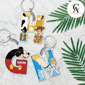 May include: Four keychains with silver rings. The keychains are shaped like the letters G, H, and N, and feature cartoon characters. The letter G is red, yellow, and black with Mickey Mouse. The letter H is yellow and white with a cow print and Woody. The letter N is blue with Cinderella.