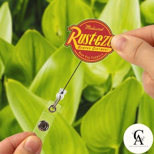 May include: A round, red and yellow "Rust-eze Bumper Ointment" badge on a retractable reel. The badge has the text "Medicated", "Bumper Ointment", and "Rear End Formula". The background is a blurred green.