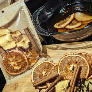 May include: Dried orange and apple slices with cinnamon sticks and cloves, arranged on a wooden cutting board. Some slices are in a clear glass dish with liquid. A clear bag contains more dried fruit.