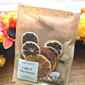 May include: A clear plastic bag filled with dried orange and lemon slices, and herbs. The bag has a small white label that reads "SHININ' SWEETS Cold & Flu Season SIMMER POT". The bag is sealed with a zipper closure. The background includes a sunflower.