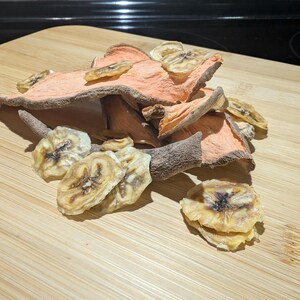 May include: A pile of dried banana and sweet potato slices on a wooden cutting board. The sweet potato slices are a light orange color, and the banana slices are a light yellow with brown spots. The cutting board has a light brown color.