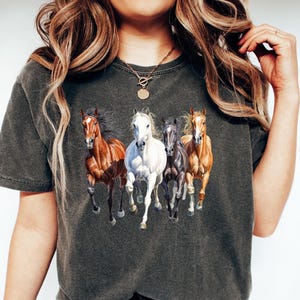 May include: Dark gray t-shirt featuring a graphic of five running horses in shades of brown, white, and gray. The horses are depicted with flowing manes and tails, creating a dynamic and energetic design. The shirt is made of a soft material.