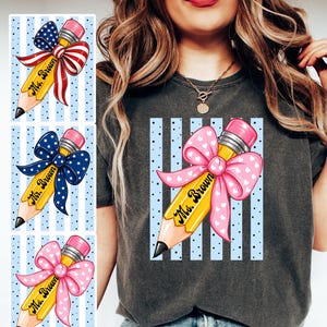 Puede incluir: Una camiseta gris carbón con un gráfico de un lápiz amarillo con goma rosa y un gran lazo rosa con corazones blancos. El lápiz lleva impreso el texto Ms. Brown sobre un fondo de rayas verticales azules y puntos pequeños.