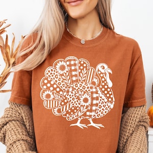 Puede incluir: Una camiseta de manga corta color óxido con un gran gráfico blanco de un pavo. El diseño del pavo está compuesto por varios patrones que incluyen girasoles, cuadros vichy, lunares y estampado de leopardo en las plumas de la cola.
