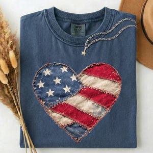 May include: A navy blue t-shirt with a heart-shaped American flag design. The heart is made of patches of blue fabric with white stars and red and white striped fabric. A silver necklace with a star pendant rests on the shirt.