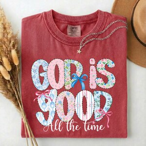 Puede incluir: Una camiseta roja con la frase "GOD IS GOOD" en un dise&ntilde;o de patchwork con estampados florales y a rayas. La frase "All the time" est&aacute; debajo. Un collar plateado con un colgante de estrella est&aacute; sobre la camiseta.